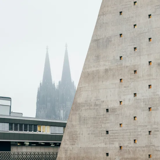 Blick auf die Oper Köln am Offenbachplatz mit ihrer markanten Betonfassade und den kleinen quadratischen Fenstern. Im Hintergrund ragen die Türme des Kölner Doms leicht im Nebel auf. Die Aufnahme zeigt das Zusammenspiel moderner Architektur und der historischen Stadtsilhouette.